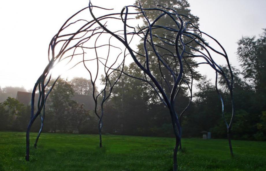 Organic steel gazebo in black made to look like branches growing from the ground, standing on a green lawn with shrubbery and trees behind, silohetted by the sun